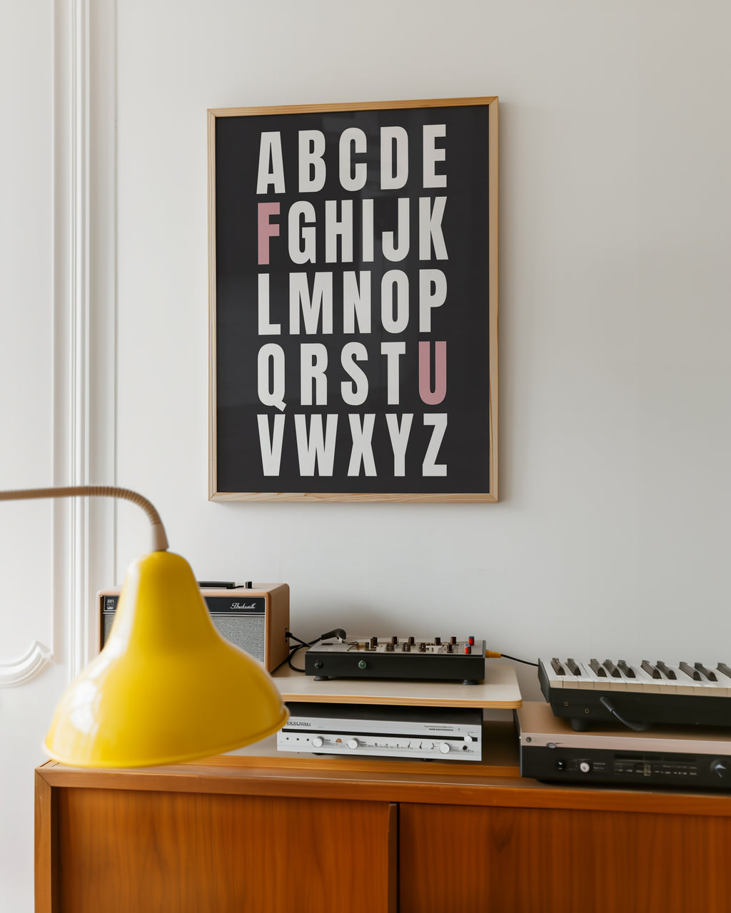 Framed alphabet poster with the letters F and U highlighted in pink on a wall above vintage electronic equipment with a yellow lamp.