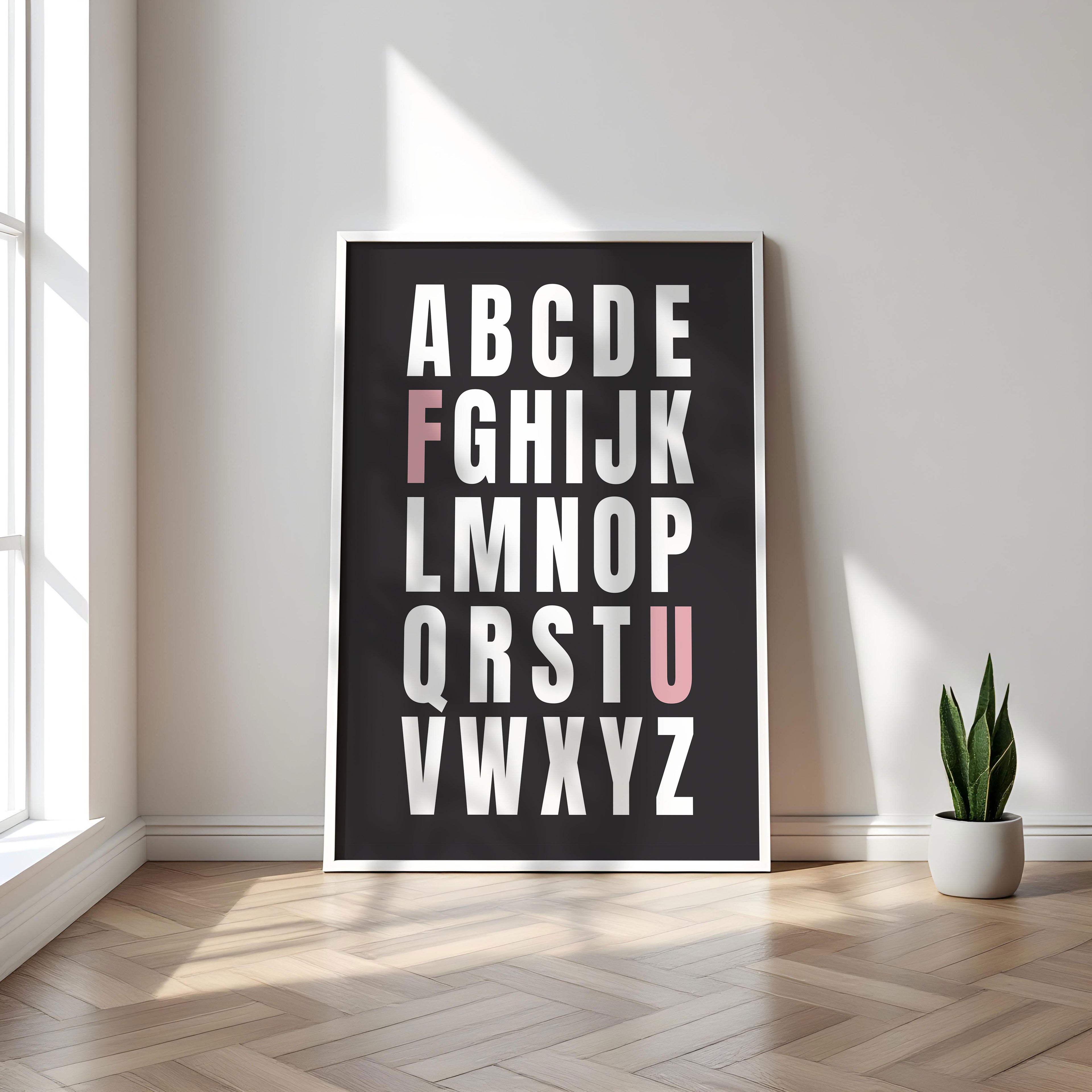 Framed alphabet poster with the letters F and U highlighted in pink on a wall with a plant on a wooden floor.
