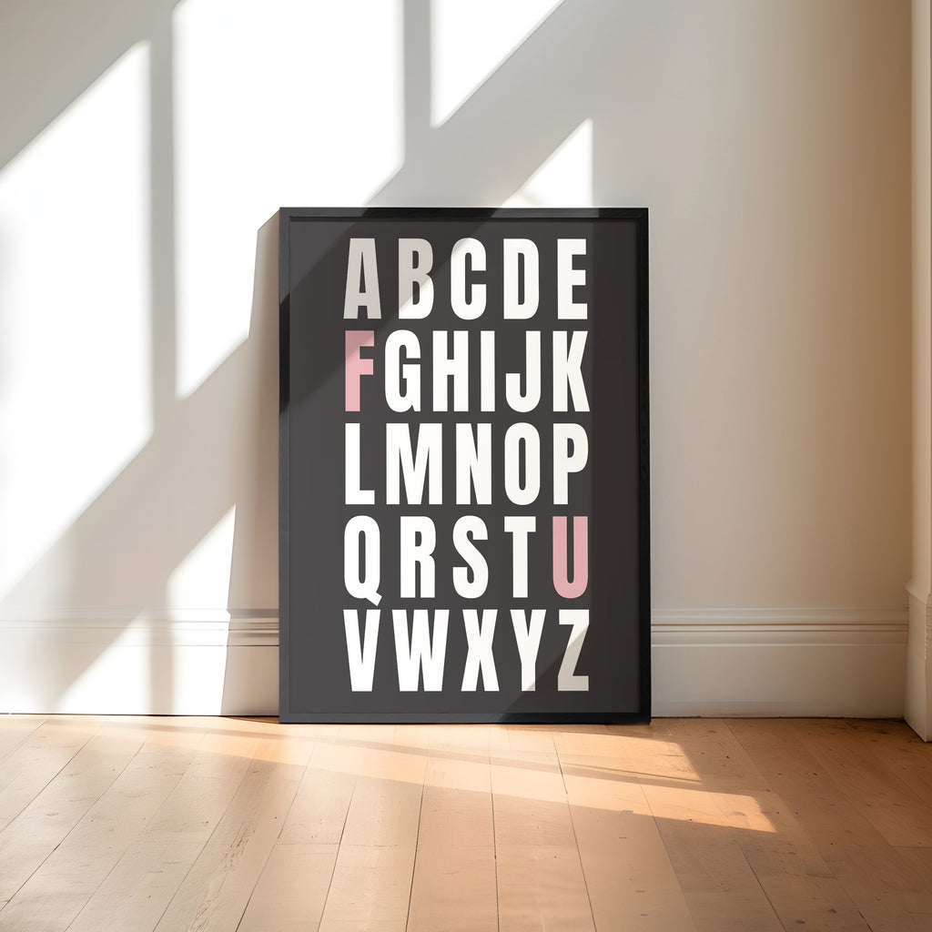 Framed alphabet poster with the letters F and U highlighted in pink on a wall with sunlight casting shadows