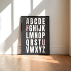 Framed alphabet poster with the letters F and U highlighted in pink on a wall with sunlight casting shadows