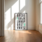 Framed alphabet poster with the letters F and U highlighted in teal on a wall with sunlight casting shadows.