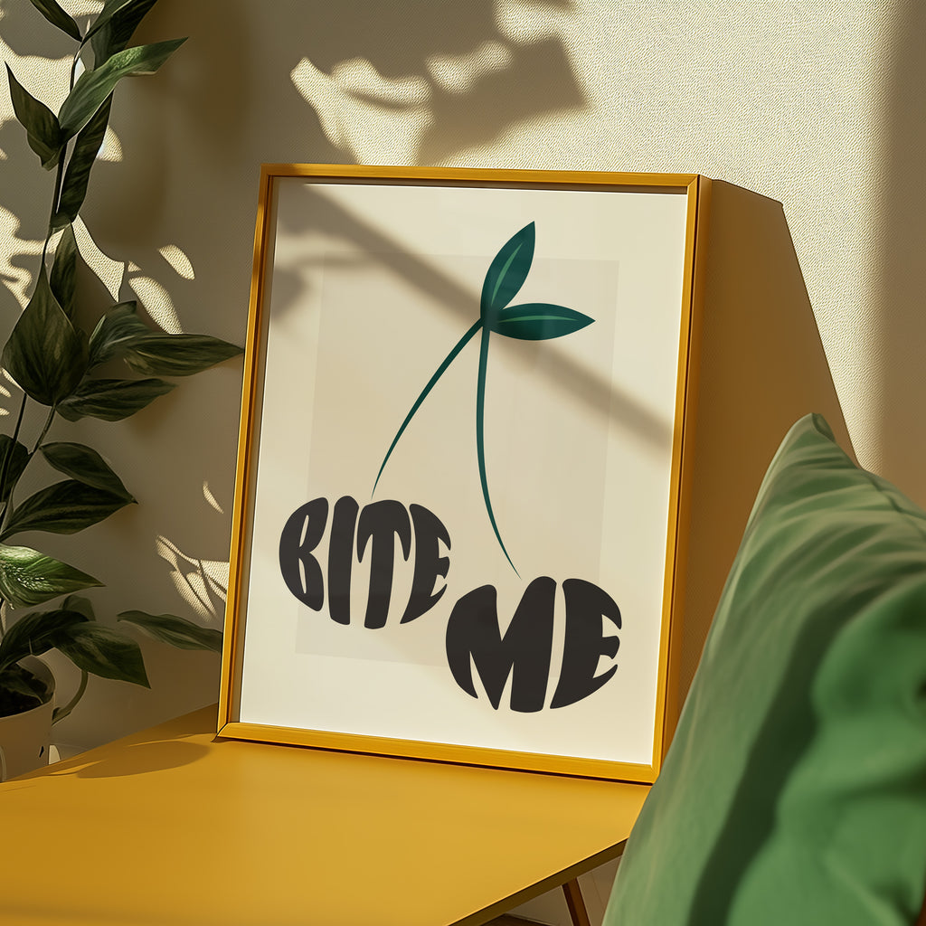 Framed artwork with 'BITE ME' text in the shape of two cherries on a yellow surface with a plant and a chair in the background.