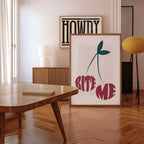 Framed artwork with 'Bite Me' text in the shape of two cherries in a room with a wooden floor and table.