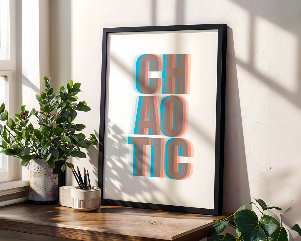 Framed artwork with the word 'CHAOTIC' in a 3D effect on a wall with plants and a window.