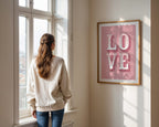 Woman standing by a window looking outside, with a 'LOVE' framed picture on the wall.