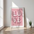 Framed poster with 'LOVE' on a pink background in a bright room with a plant.