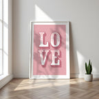 Framed poster with 'LOVE' on a pink background in a bright room with a plant.