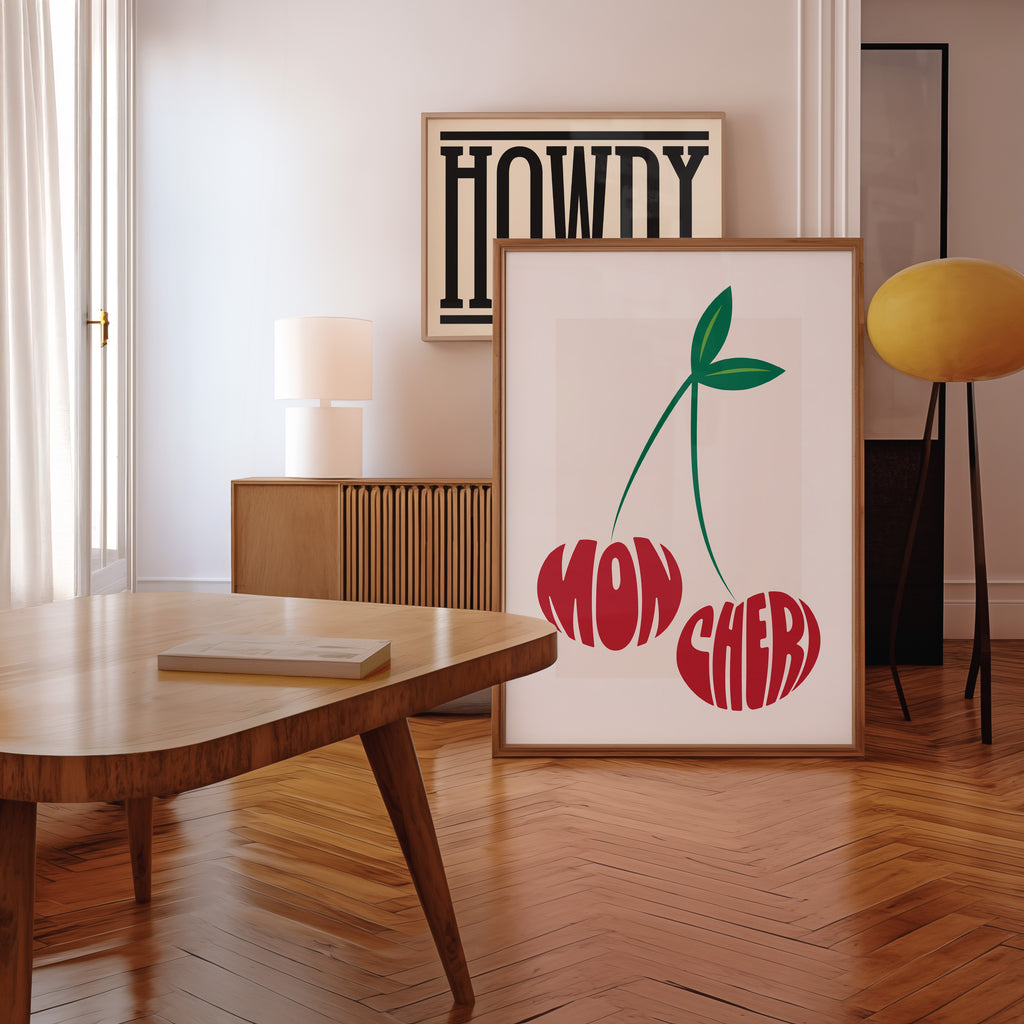 Framed artwork with 'Mon Cheri' text in the shape of two cherries in a room with a wooden floor and table.