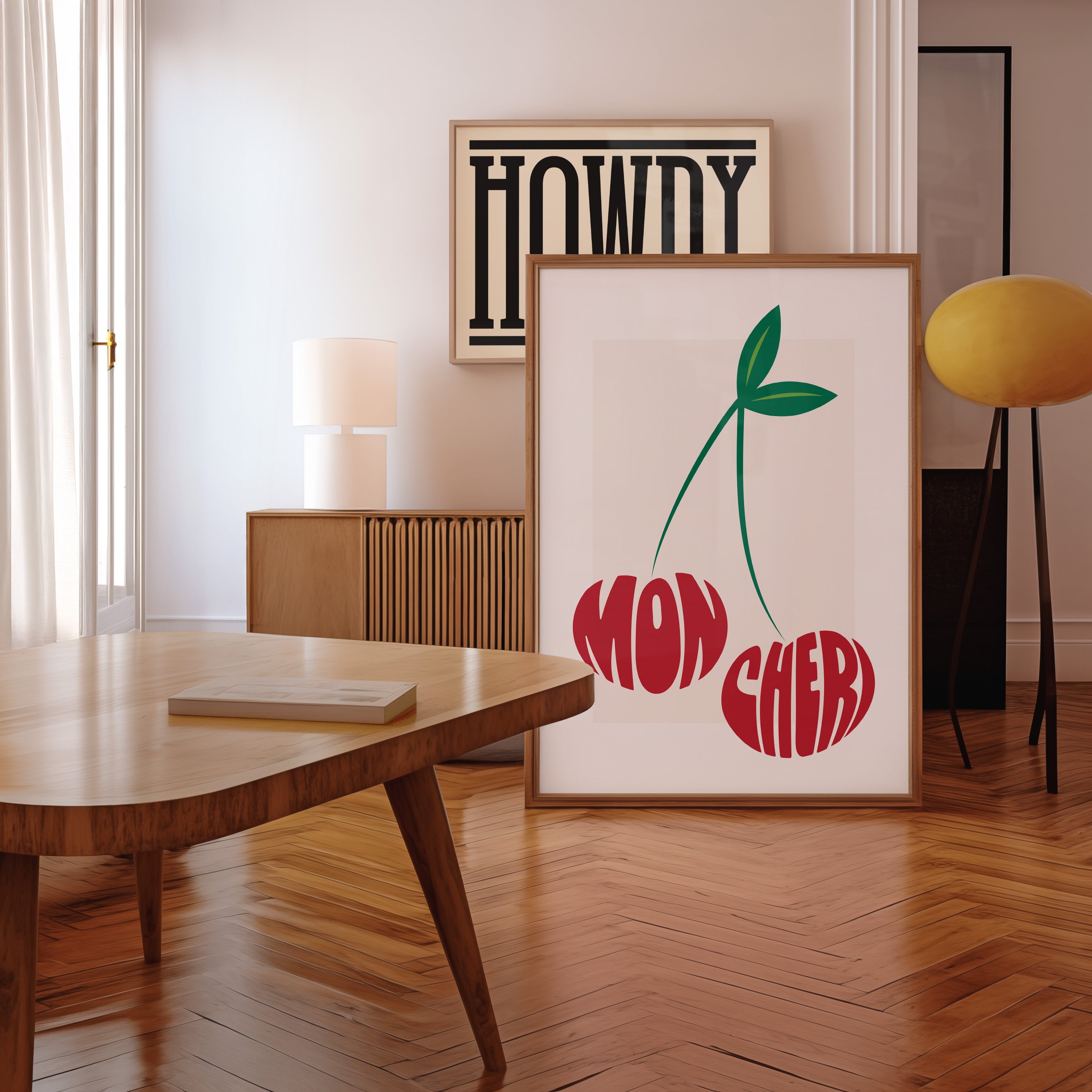 Framed artwork with 'Mon Cheri' text in the shape of two cherries in a room with a wooden floor and table.