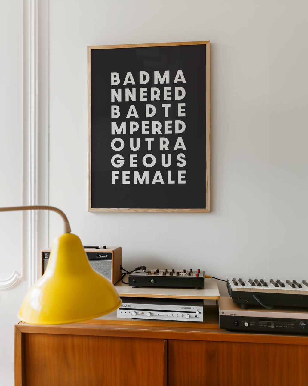 Framed poster with text saying 'Bad Mannered, Bad Tempered, Outrageous Female' on a wall above vintage electronic equipment.