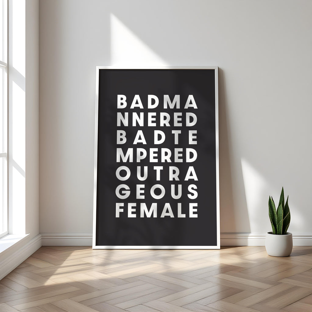 Framed poster with text saying 'Bad Mannered, Bad Tempered, Outrageous Female' on a wall in a room with a plant.