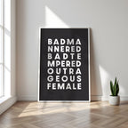 Framed poster with text saying 'Bad Mannered, Bad Tempered, Outrageous Female' on a wall in a room with a plant.