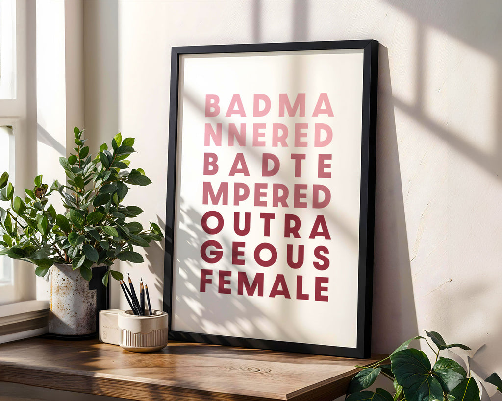 Framed poster saying 'Bad Mannered, Bad Tempered, Outrageous Female' on a wall next to plants