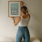 A person hanging a framed poster saying 'Bad Mannered, Bad Tempered, Outrageous Female'.