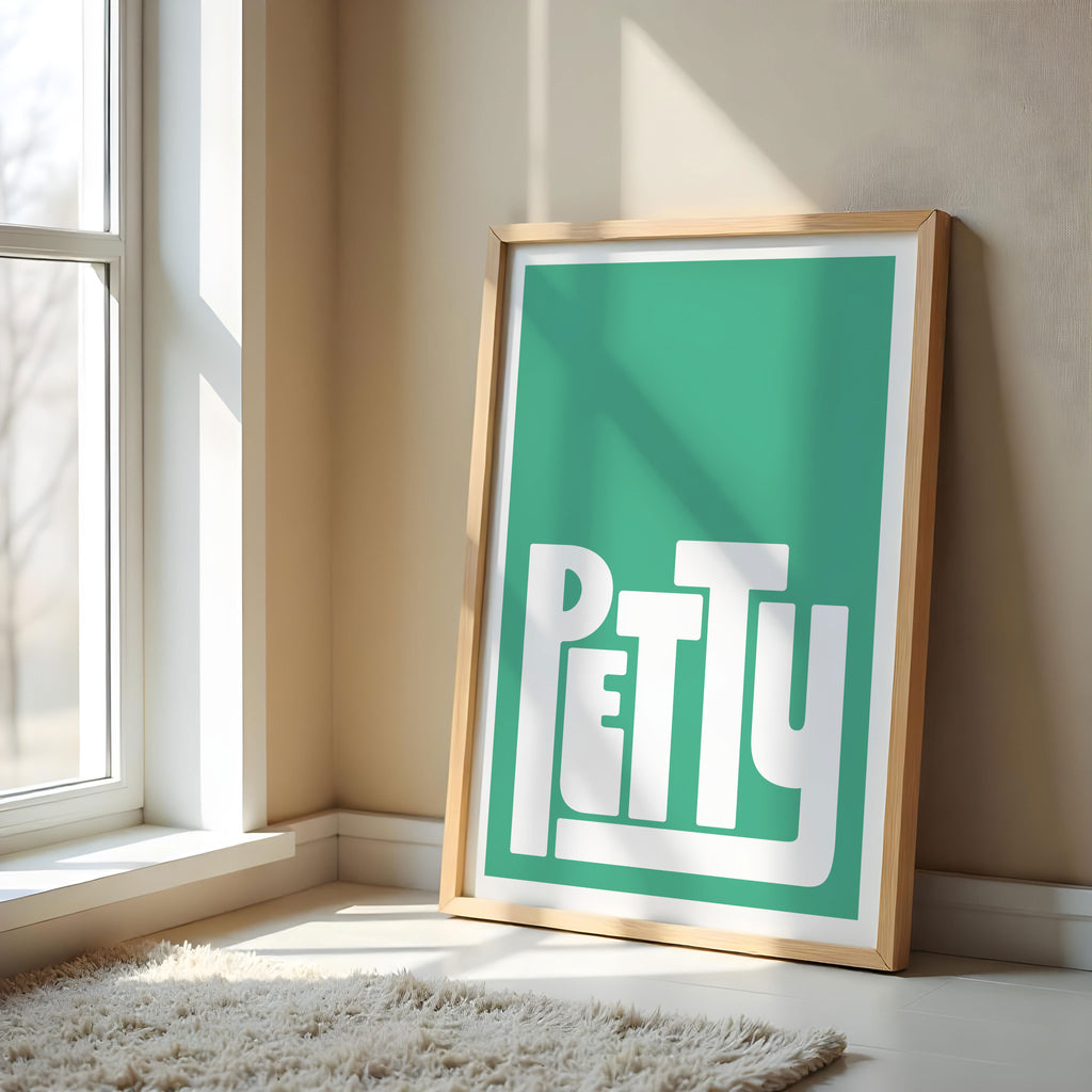 Framed artwork with the word 'PETTY' on a teal background, leaning against a wall near a window.