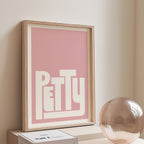 Framed artwork with 'PETTY' on a pink background, placed on a shelf against a beige wall.