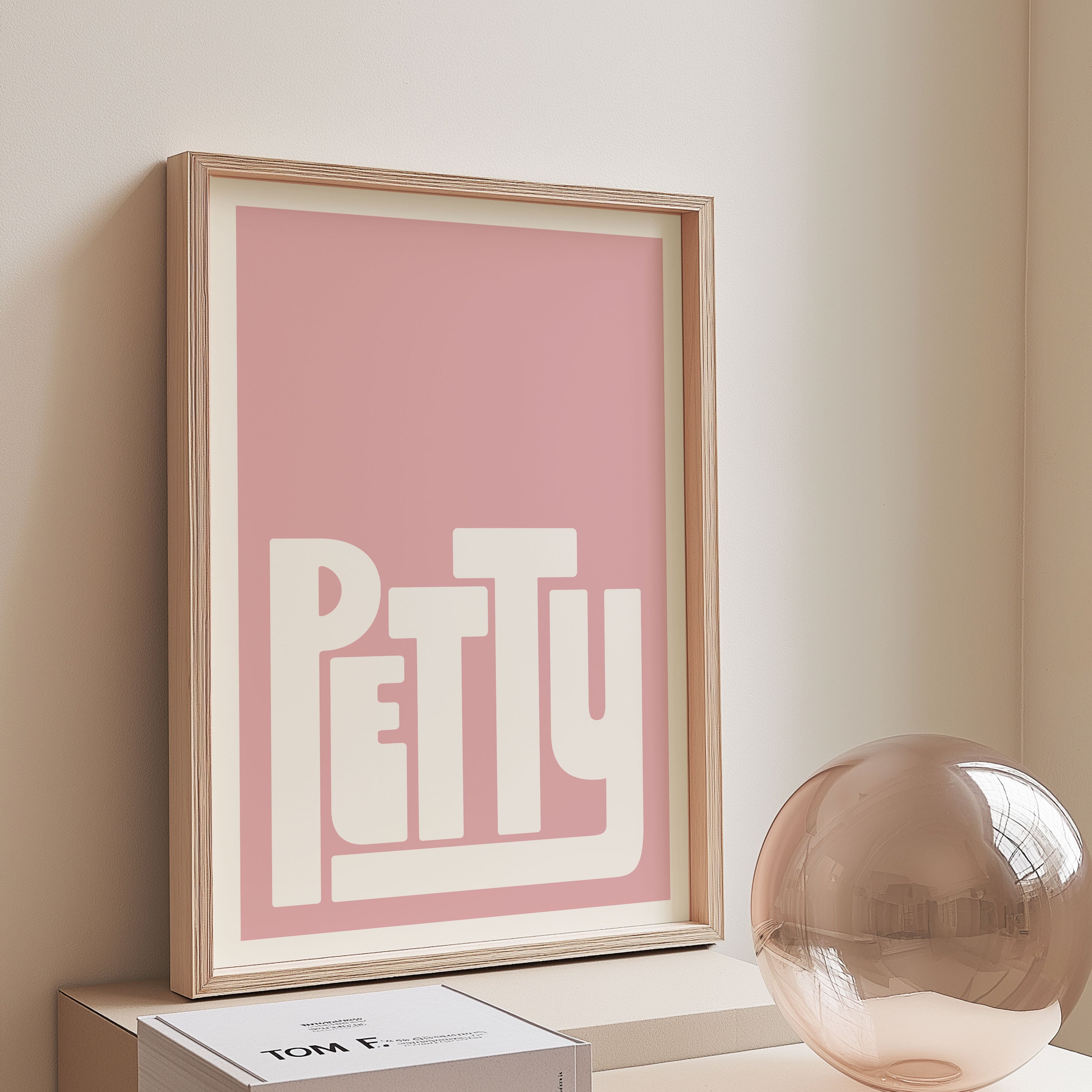 Framed artwork with 'PETTY' on a pink background, placed on a shelf against a beige wall.