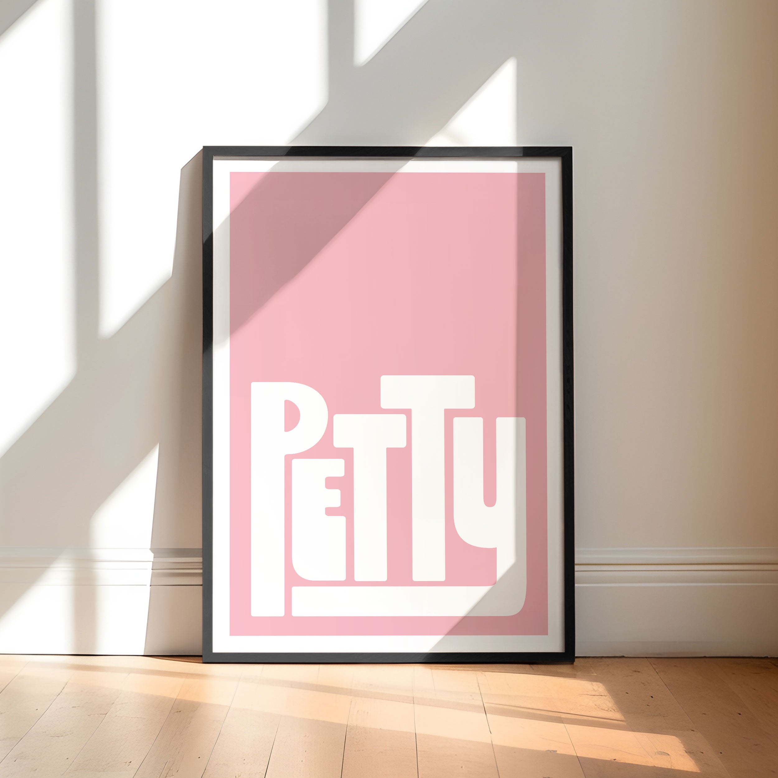 Framed poster with 'PETTY' text on a pink background, placed against a wall with sunlight casting shadows.